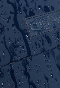 Tejido impermeable en color azul marino con gotas en la superficie, que presenta un distintivo parche con el logo en relieve de Eastpak y una textura suave.