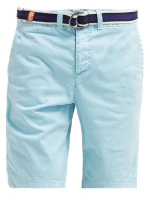 Mand iført sort polo-shirt, lyseblå shorts og sorte sneakers, holder sorte solbriller i højre hånd, står mod en ensfarvet baggrund.