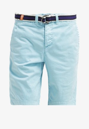 Mężczyzna ubrany w czarną koszulkę polo, jasnoniebieskie szorty i czarne trampki, trzymający czarne okulary przeciwsłoneczne w prawej ręce, stojący na jednolitym tle.