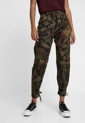 Osoba ubrana w moro spodnie typu cargo z kieszeniami na guziki oraz czarne trampki z białą podeszwą, stojąca na jednolitym tle.