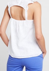 Top bianco senza maniche con motivo floreale testurizzato, scollatura arricciata e design della schiena aperta, abbinato a pantaloni blu.