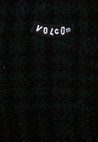 Gebreide trui in donkergroen en zwart met een gestructureerde stof. Voorzien van een opvallend zwart label met wit "VOLCOM" tekstlogo.