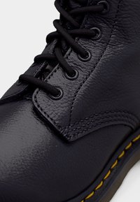Botte en cuir noir avec un design à tige haute, des lacets ronds, des œillets métalliques, des coutures jaunes et une finition texturée sur la surface.