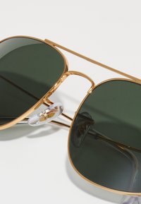 Occhiali da sole in metallo color oro con grandi lenti verde scuro. Le caratteristiche includono naselli trasparenti e un design a doppio ponte.