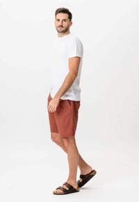 Mand står iført en hvid t-shirt, rustfarvede shorts og mørkebrune sandaler mod en helt hvid baggrund.