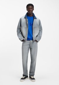 Chaqueta de mezclilla clara con cuello azul, combinada con una camiseta gráfica azul y jeans grises. Zapatillas negras con suelas blancas completan el conjunto.