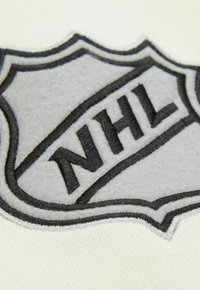 Ein weißer Stoff mit einem gestickten NHL-Logo in Grau und Schwarz, das in Form eines Wappens dargestellt ist, mit detailliertem Stich und Textur.