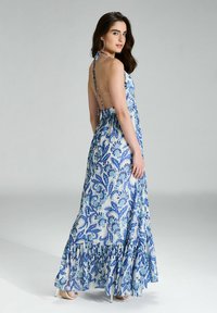 Robe maxi avec un motif floral bleu et blanc, décolleté halter, corsage ajusté et ourlet évasé à volants. Tissu léger avec un design fluide.
