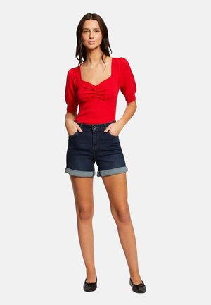 Haut rouge ajusté avec des manches bouffantes, doté d'un devant froncé. Associé à un short en denim noir, avec un ourlet retroussé. Simples ballerines noires.