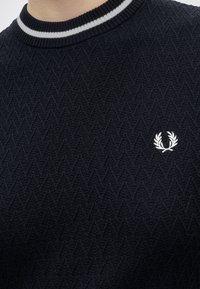 Pull texturé bleu marine avec logo couronne de laurier blanc sur la poitrine et col côtelé avec rayures blanches et bleu marine.
