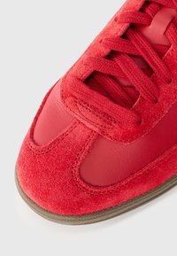 Baskets rouges en daim et cuir, avec des lacets texturés et un bout arrondi. Semelle légèrement rembourrée avec coutures visibles.