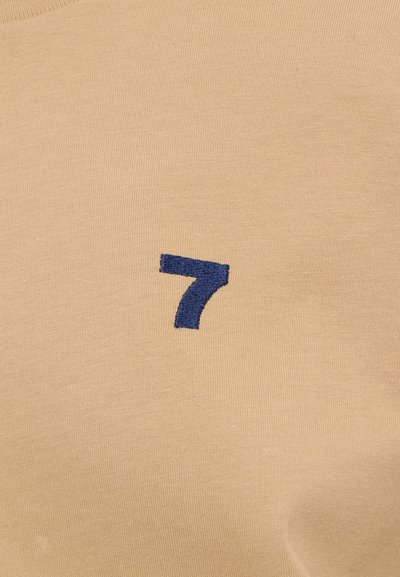 Beige cotton fabric with a soft texture featuring a dark blue embroidered number "7" on the front. Simple, minimal design.