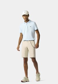 Lichtblauw poloshirt met een klein geborduurd logo, beige shorts met een kettingdetail en lichtgekleurde sneakers. Witte pet gedragen.