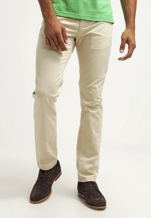 Uomo che indossa pantaloni beige slim fit, scarpe stringate marrone scuro e una camicia verde, in piedi davanti a uno sfondo bianco uniforme.