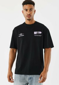 Camiseta de algodón negra con mangas cortas, de corte oversize, con estampados gráficos en blanco y rosa claro en la parte delantera y en el lado izquierdo del pecho.