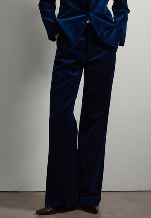 Pantalones de traje de terciopelo azul marino con un diseño de pierna ancha, con una textura suave y un ajuste entallado, combinados con zapatos de un marrón más oscuro.