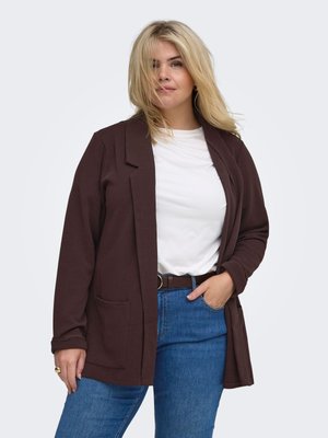 Blonde vrouw met wit hemd, donkerbruine blazer en blauwe jeans, staand met één hand in de zak tegen een effen achtergrond.