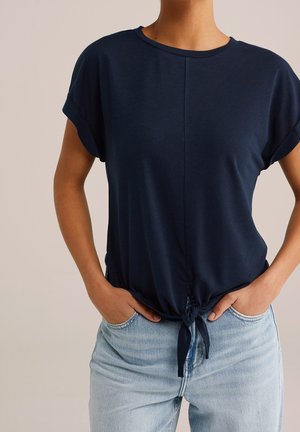 Kvinde iført en marineblå kortærmet top med knude foran og lyseblå højtsiddende jeans, hænderne i lommerne.