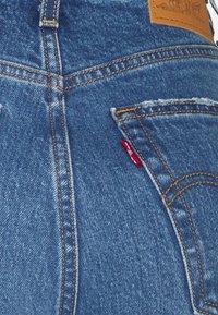 Jeans de mezclilla azul con costuras naranjas, una cintura alta y una pequeña etiqueta roja en el bolsillo trasero. Detalles desgastados cerca de los bordes de los bolsillos.