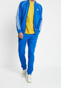 Blå träningsoverall i lätt tyg, med vita ränder längs ärmarna och sidorna, samt en gul t-shirt under.