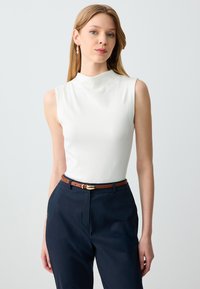 Weißes ärmelloses Oberteil mit hoher Halslinie, kombiniert mit navyfarbener Hose und einem braunen Gürtel mit goldener Schnalle. Glatte Textur und taillierte Passform.