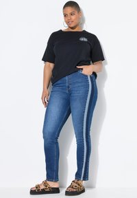 Mustat puuvillapaidat, joissa on pieni graafinen printti, vaaleansiniset farkut, joissa on vaaleansiniset sivu raidat, ja ruskeat leopardikuvioiset sandaalit paksulla pohjalla.