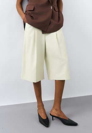 Osoba v voľných, krémových šortkách po kolená, hnedom bezrukávovom saku a čiernych lodičkách na podpätku so zašpicatenou špičkou. Ruky v vreckách.