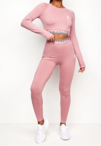 Růžový dlouhý rukáv crop top a odpovídající legíny s vysokým pasem, vyrobené z hladké látky, s logem na pásu a bezešvým designem.