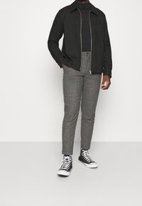 Schwarz-weiß gestreifte Zip-Jacke über einem schwarzen Rollkragenpullover, kombiniert mit grauen strukturierten Hosen und schwarzen High-Top-Sneakern.