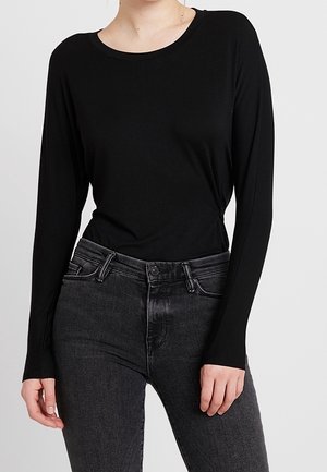 Femme portant un haut noir ajusté à manches longues rentré dans un jean taille haute gris foncé, debout avec une main détendue le long du corps.