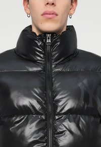 Jachetă puffer neagră cu guler înalt, finisaj lucios, fermoar frontal și detaliu subtil al logo-ului pe piept. Textură netedă și design matlasat.