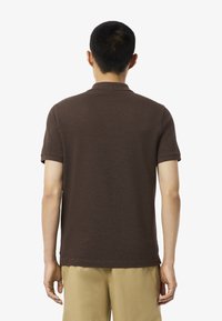 Henkilö seisoo kameraan selkä kohti, päällään ruskea polo paita ja khaki-housut, esitellen yksinkertaista ja rentoa asua.