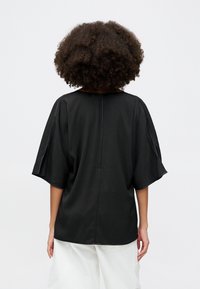 Sort løs pasform top med korte ærmer, struktureret halsudskæring og en glat tekstur. Sømmen løber vertikalt ned ad ryggen.