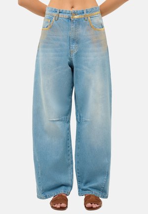 Personne portant un jean en denim bleu clair ample et des sandales marron ouvertes aux orteils, visible de la taille aux pieds sur un fond uni.