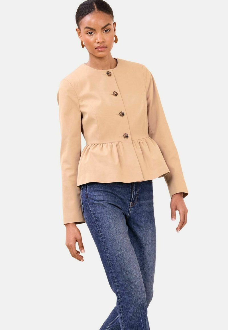 Sieviete, tērpusies bēša krāsas pogātu pepluma žaketi ar garām piedurknēm un zilas džinsas, stāvot pret vienkāršu gaišu fonu.