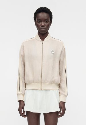 Osoba o ciemnej karnacji ubrana w beżową kurtkę na zamek błyskawiczny z czarnymi paskami na rękawach i małym okrągłym logo, w połączeniu z białą plisowaną spódnicą.