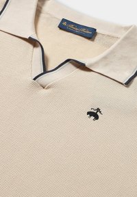 Beige Polo-Shirt aus strukturiertem Stoff, mit V-Ausschnitt und Kragen in Marineblau. Mit einem kleinen schwarzen gestickten Logo auf der Brust.