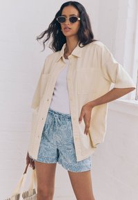 Camicia oversize beige chiaro con maniche corte, canotta bianca e pantaloncini blu con motivo a foglie in rilievo. La modella tiene in mano una borsa a righe.