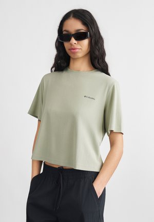 Γυναίκα με μακριά μαλλιά που φοράει μαύρα γυαλιά ηλίου, ανοιχτό πράσινο κοντομάνικο τοπ και μαύρο παντελόνι με κορδόνι, τα χέρια στις τσέπες, ουδέτερο φόντο.