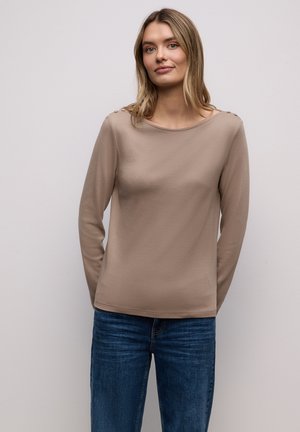 Haut beige à manches longues en tissu doux avec un col rond, doté de trois boutons sur chaque épaule, associé à un jean bleu.