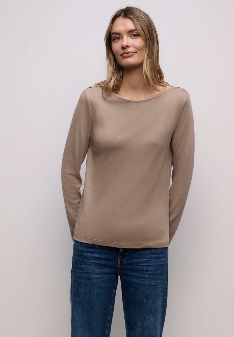 Top beige de manga larga hecho de tela suave con escote redondo, con tres botones en cada hombro, combinado con jeans azules.
