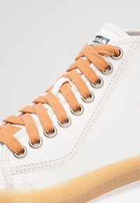 Białe płócienne sneakersy z pomarańczowymi sznurówkami, metalowymi oczkami i jasnobrązową gumową podeszwą, ze zbliżeniem na bok i logo na języku.