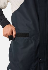Salopette imperméable bleu marine en tissu texturé, dotée d'une poche avec velcro et d'accents de boutons-pression sur le côté.
