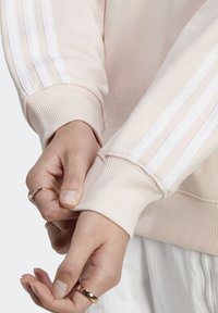 Světle růžová mikina s bílým designem tří pruhů na rukávech, žebrovanými manžetami a jemnou texturou. Ruce upravují látku rukávu.