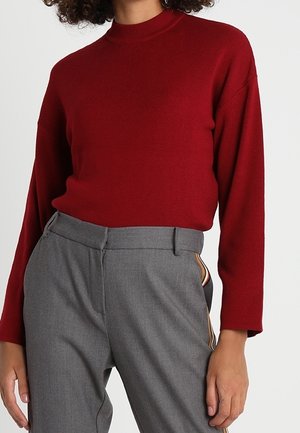 Femme portant un haut rouge ajusté à manches longues, rentré dans un pantalon gris taille haute avec un détail de bande latérale.