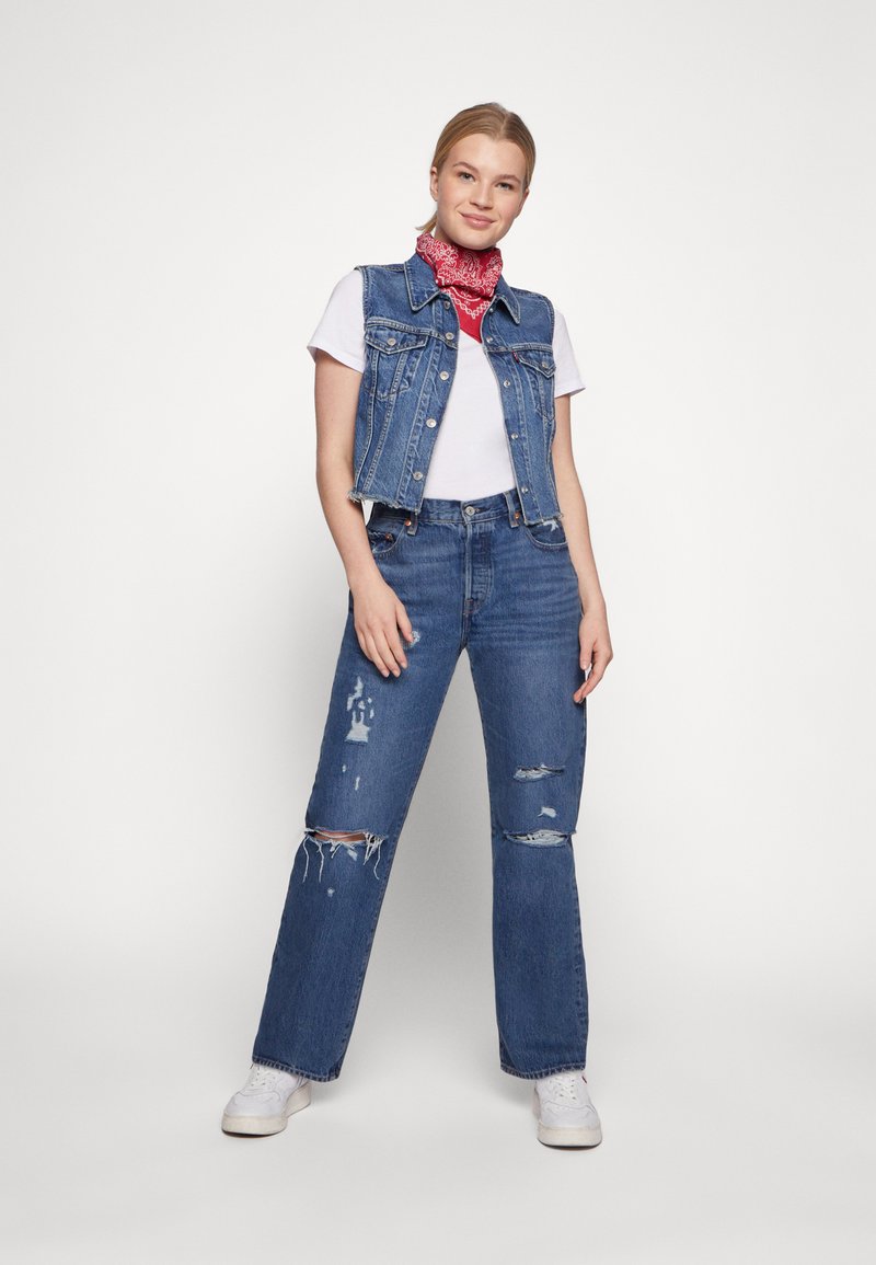 Mujer joven vistiendo jeans desgastados de color azul, chaleco de denim sobre una camiseta blanca, bufanda de cuello con un patrón rojo y zapatillas blancas, de pie sobre un fondo blanco.