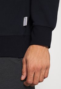 Námořnický modrý svetr s manžetami a spodním lemem z žebrovaného materiálu, na levé straně s malou textilní visačkou s nápisem "Scotch & Soda". Měkčená textura.