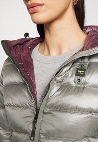 Doudoune gris métallisé avec doublure intérieure violette, dotée d'une fermeture éclair, d'une capuche et d'une écusson logo sur la poitrine.