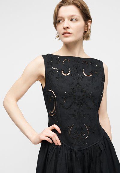 Kobieta o krótkich włosach w czarnej, bezrękawowej sukience z misternym haftem w kształcie kwiatów, opierająca rękę na biodrze, patrząca w bok.