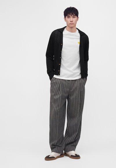 Giovane uomo che indossa un cardigan nero abbottonato sopra una t-shirt bianca, pantaloni a righe a gamba larga e sneakers bianche e marroni, in piedi con le mani in tasca.